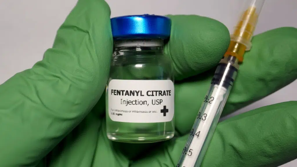 Hand wearing a green glove holding a vial of fentanyl citrate and a needle. Fentanyl has a high risk of overdose.