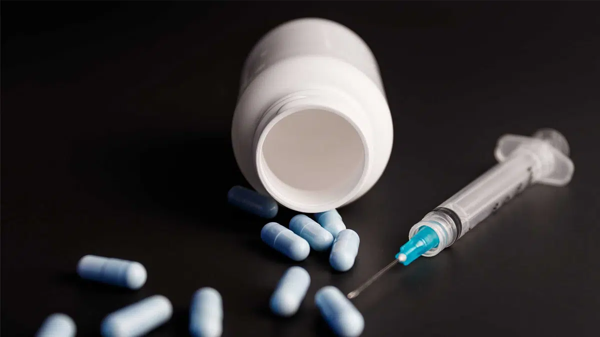 White pill bottle with blue and white pills spilling out and a blue and white needle. Synthetic opioids have diverse chemical compositions.