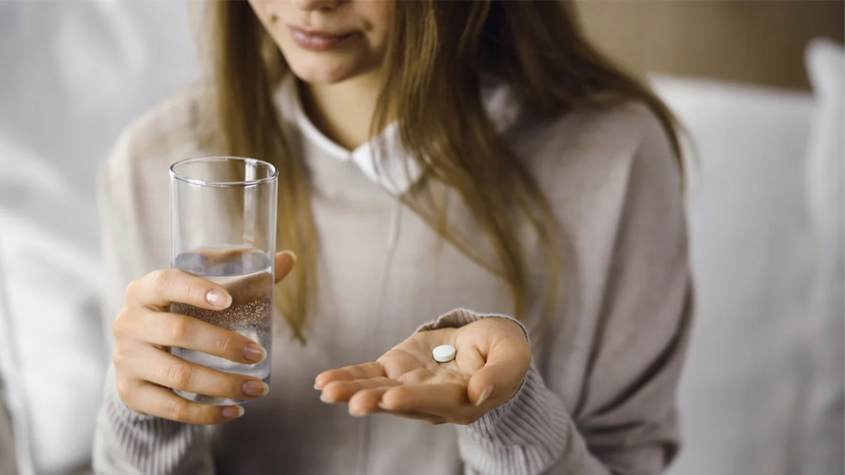 Woman holding a white pill in one hand and a glass of water in the other.
