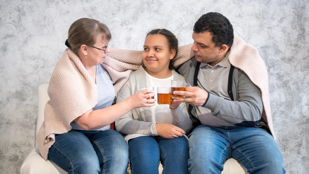 A family of three sitting on a couch, sharing a warm moment. The parents are wrapped in a light pink knitted blanket and holding glasses of tea, while their child sits between them, smiling.