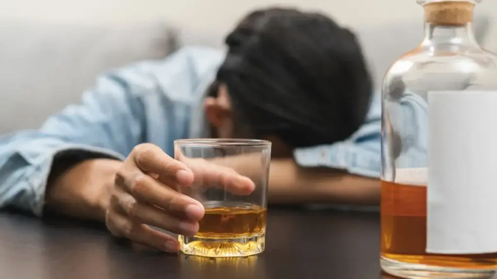A person with dark hair wearing a denim shirt rests their head on a table, appearing intoxicated. Their hand holds a glass of whiskey, and a bottle of alcohol with a blank label sits nearby.