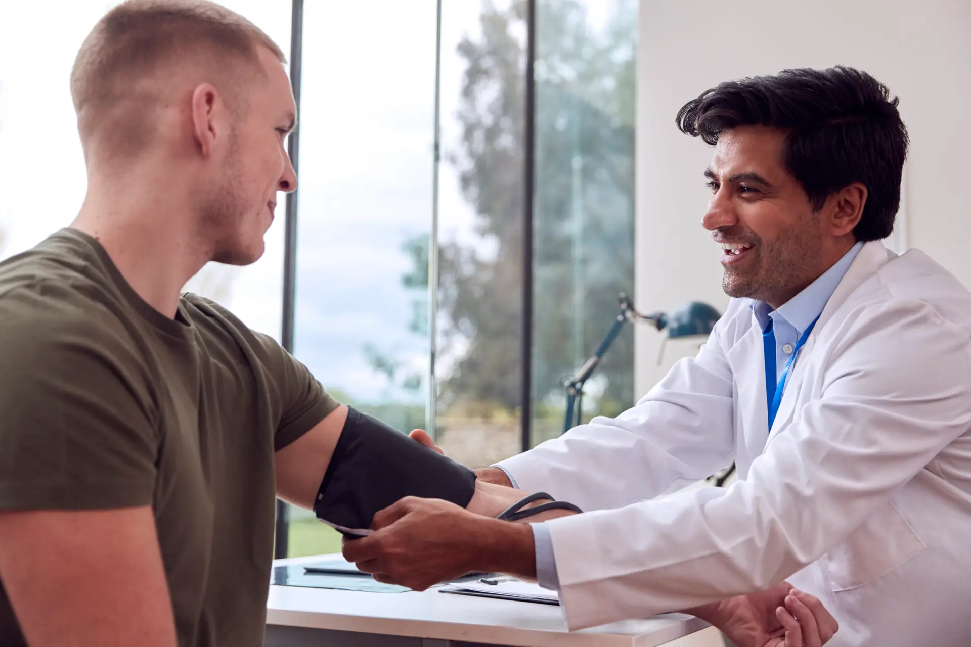 Male Doctor Or GP Examining Young Male Patient Taking Blood Pressure With Sphygmomanometer