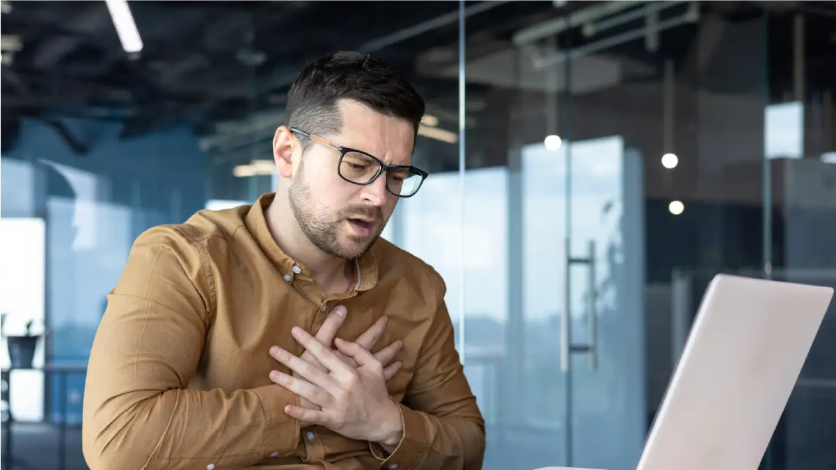 A man wearing glasses and a brown shirt sits in an office setting, clutching his chest with both hands and looking distressed.