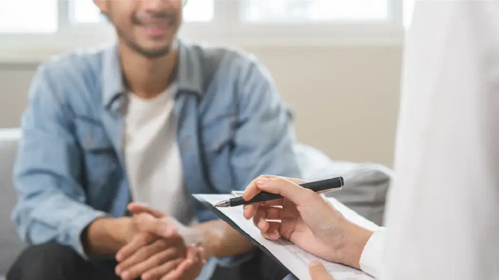 A smiling man wearing a denim jacket sits across from a therapist, who is holding a clipboard and pen, in a bright and professional setting.