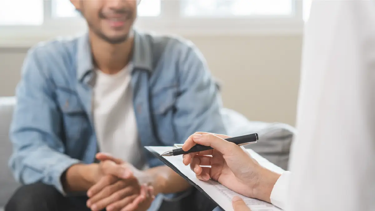 A smiling man wearing a denim jacket sits across from a therapist, who is holding a clipboard and pen, in a bright and professional setting.