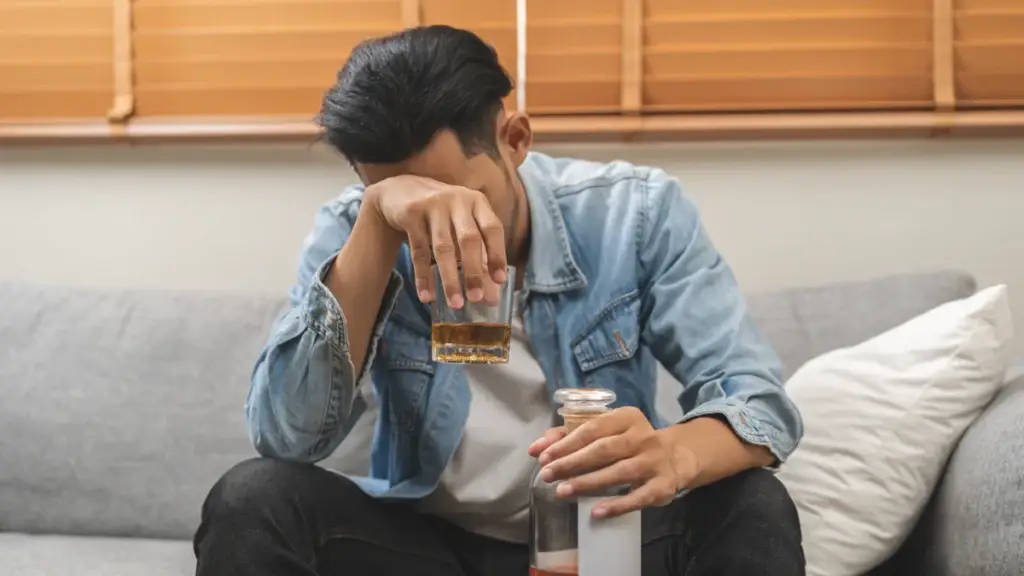 A man is sitting on a couch, wearing a denim jacket, holding a glass of liquor in one hand and a bottle in the other. His posture and expression suggest emotional distress or contemplation.