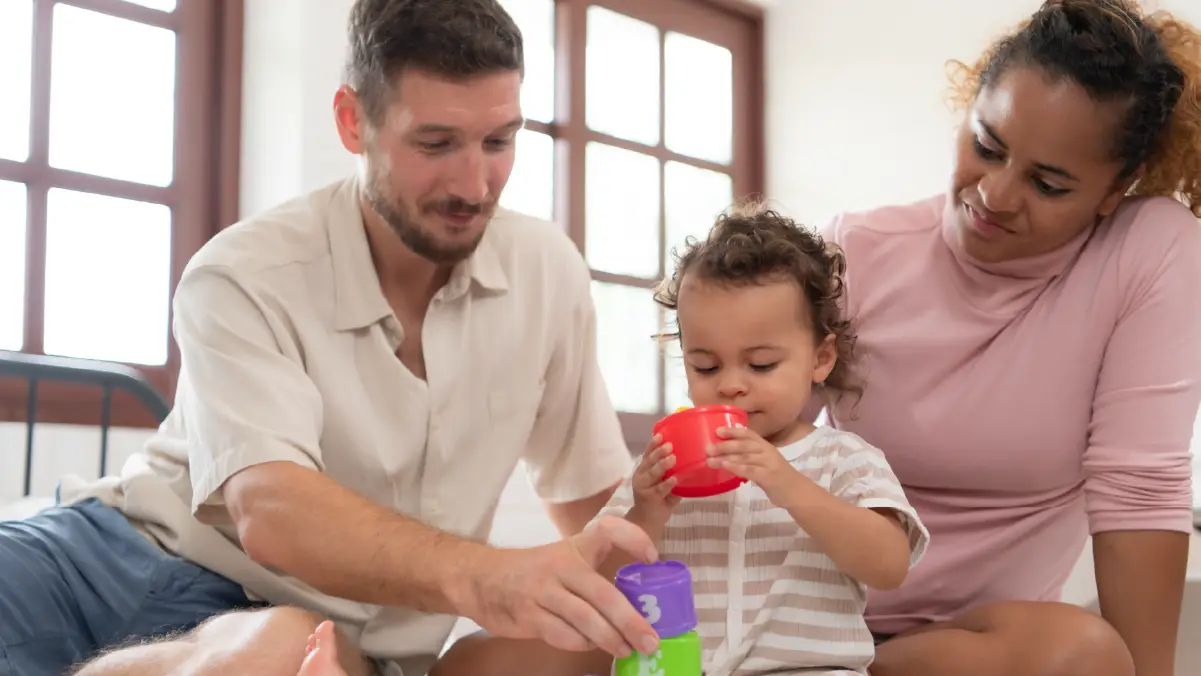 young family playing together indoors, with a father stacking cups, a toddler engaging with toys, and a smiling mother watching.