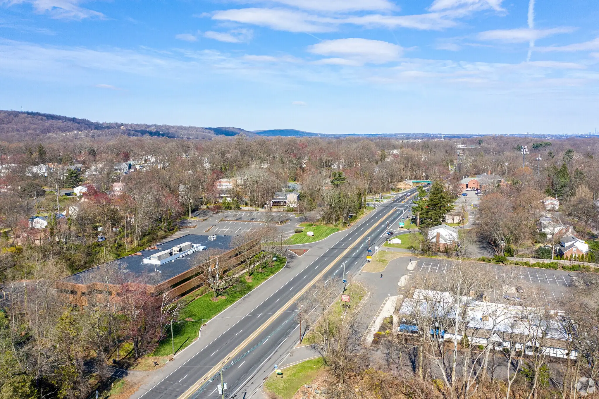 Aerial view of a suburban roadway surrounded by trees, parking lots, and low-rise buildings, with rolling hills visible in the distance under a blue sky.