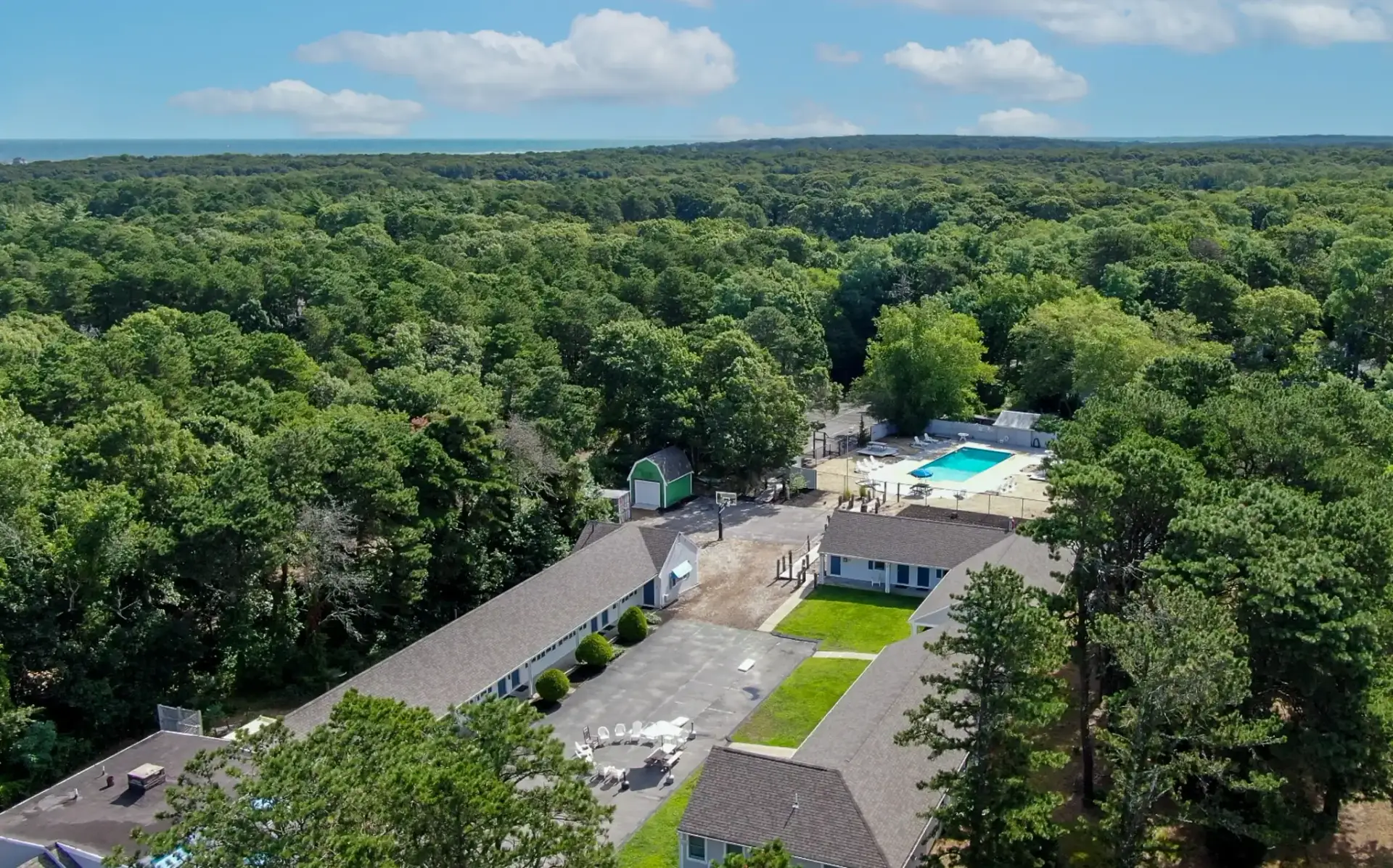 Aerial view of a residential-style campus surrounded by dense forest, featuring several low buildings, a central courtyard, and an outdoor swimming pool under a clear blue sky.