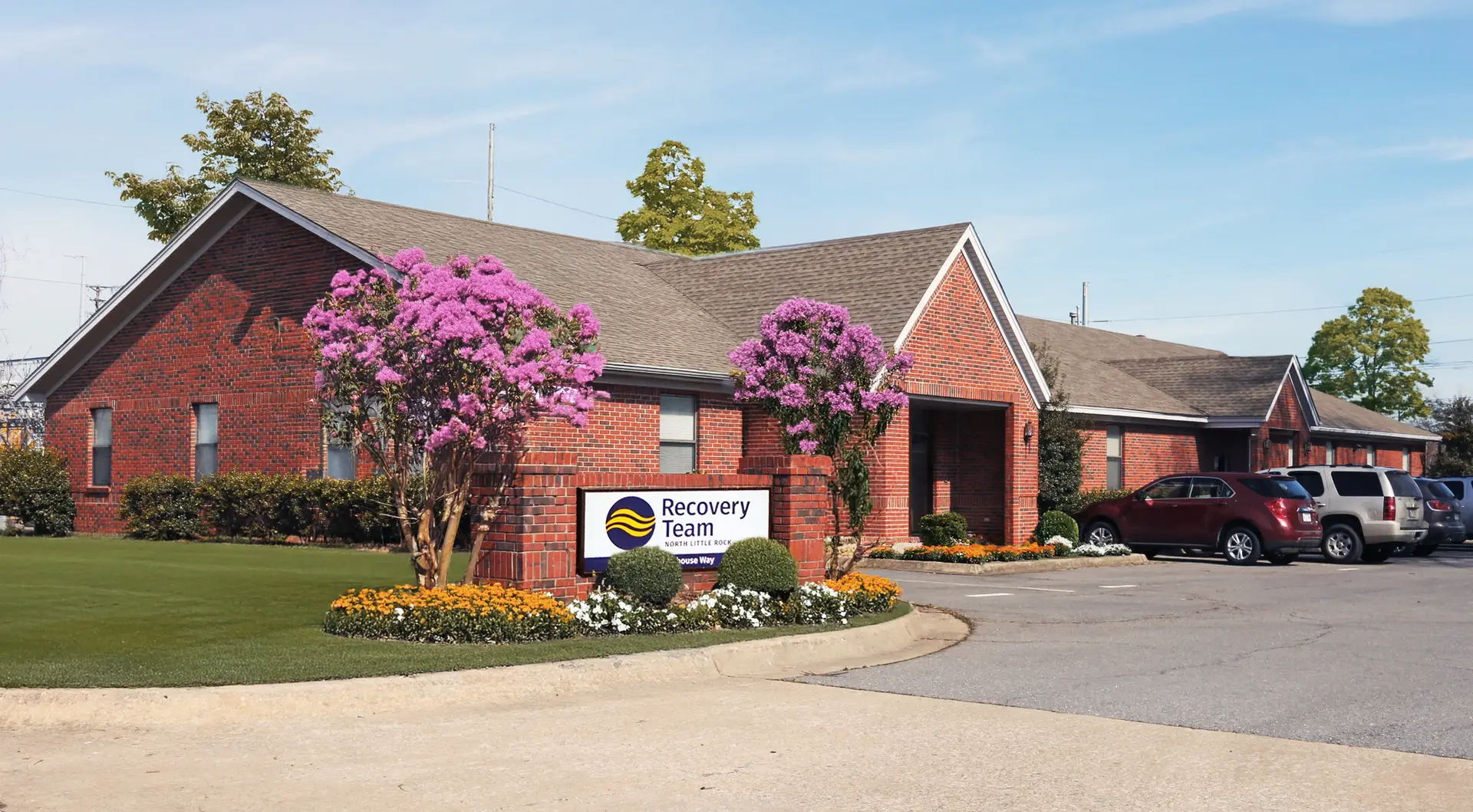 A two-story brick building with a "Recovery Team" sign and a parking lot, surrounded by green grass and flowering bushes.