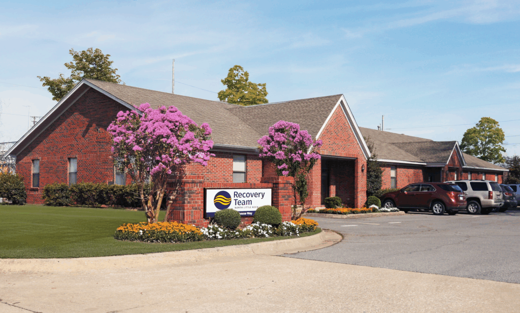 Exterior view of The Recovery Team North Little Rock treatment center in Arkansas, showing the brick building, landscaped flowers, and parking area under a clear sky.