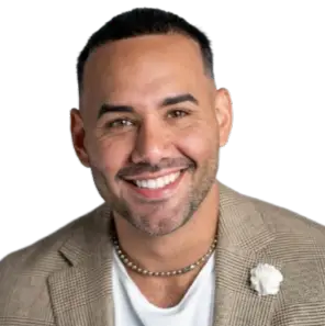 Smiling man wearing a tan blazer over a white shirt with a lapel flower pin, photographed against a dark background.